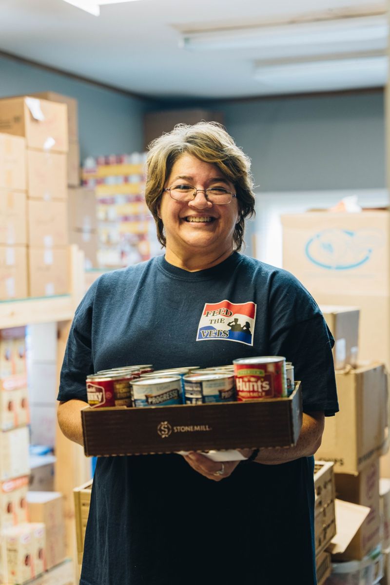 A smiling Feed the Vets volunteer holding a tray of groceries inside the pantry